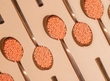 Mehrere glänzende, metallische Löffel, die diagonal von unten links nach oben rechts über eine beige Oberfläche angeordnet sind. Jeder Löffel ist gefüllt mit kleinen, roten Linsen.