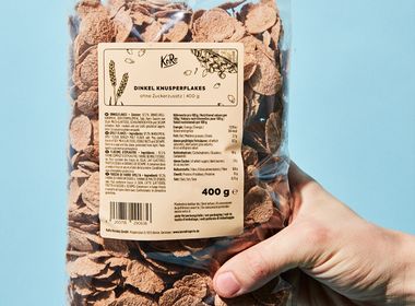 Eine Hand hält eine transparente Plastikverpackung von KoRo Dinkel Knusperflakes. Das Etikett der Verpackung zeigt die Nährwerttabelle, Zutatenliste und den Claim "Ohne Zuckerzusatz". Der Hintergrund ist hellblau.