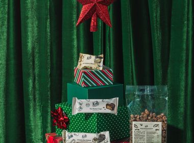 Eine festliche Weihnachtsinszenierung vor grünem Samthintergrund. Geschenkboxen in verschiedenen Grüntönen und Rot, gestapelt in Form eines Tannenbaums mit einem roten Stern als Spitze, umgeben von veganen Koro-Produkten wie Nussmus, Proteinriegeln, Nut Butter Cups und gerösteten Kichererbsen.