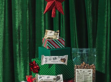 Eine festliche Weihnachtsinszenierung vor grünem Samthintergrund. Geschenkboxen in verschiedenen Grüntönen und Rot, gestapelt in Form eines Tannenbaums mit einem roten Stern als Spitze, umgeben von veganen Koro-Produkten wie Nussmus, Proteinriegeln, Nut Butter Cups und gerösteten Kichererbsen.