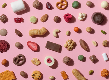  Eine vielfältige Auswahl an Snacks, darunter Nüsse, Gummibärchen, Schokolade, Brezeln und getrocknete Früchte, ist in einem gleichmäßigen Raster auf einem hellrosa Hintergrund angeordnet. 