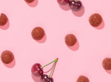 Ansicht der runden Frucht-Bites zusammen mit frischen Kirschen als dekoratives Pattern auf rosa Grund.