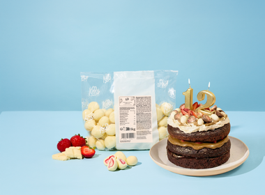 Vegane schokoladige Geburtstagstorte mit goldenen Zahlenkerzen „12“, dekoriert mit gefriergetrockneten Erdbeeren in weißer Schokolade sowie frischen Erdbeeren. Daneben steht eine große 1-kg-Tüte mit gefriergetrockneten Erdbeeren in weißer Schokolade, dazu frische Erdbeeren und Stücke weißer Schokolade vor hellblauem Hintergrund.