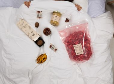 Vogelperspektive einer Person im weißen Bett, auf dessen Decke verschiedene Snacks wie gefriergetrocknete Früchte, Kekse und ein Glas Aufstrich dekorativ angerichtet sind.