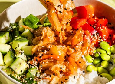 Eine bunte Bowl mit Reis, Gurken, Paprika und Edamame, aus der mit Holzstäbchen eine Portion Kimchi gehoben wird, angerichtet auf hellgrünem Grund.