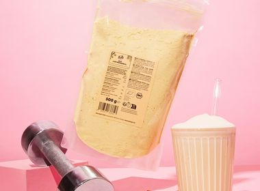Eine Hand hält eine Packung KoRo Bio Lupinenmehl neben einer Hantel und einem Protein-Shake auf rosa Hintergrund.