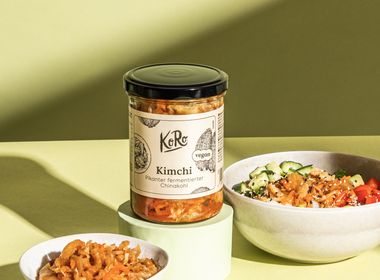 Glas mit Kimchi als Beispiel für fermentierte Lebensmittel, daneben eine Schale mit fermentiertem Kohl und eine Bowl mit frischem Gemüse auf hellgrünem Hintergrund.
