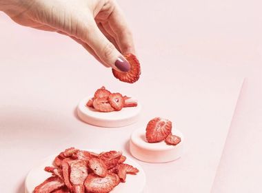 Eine Hand greift nach einer gefriergetrockneten Erdbeerscheibe. Darunter liegen weitere getrocknete Erdbeerscheiben als Snack arrangiert auf drei kleinen, rosa Podesten auf einem rosa Hintergrund.