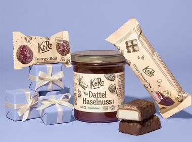 Produktset von KoRo mit einem Glas Bio Dattel-Haselnuss-Creme, einem Energy Ball Kakao & Himbeer und einem Crispy Protein Bar Peanut vor lila Hintergrund mit kleinen Geschenkpäckchen.