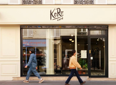 Zwei Passanten gehen an der großen Fensterfront des hellen „KoRo Café“ vorbei, das sich im Erdgeschoss eines klassischen Pariser Gebäudes befindet.