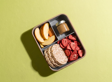 Draufsicht auf eine strukturierte Bento-Box im Querformat auf hellgrünem Hintergrund, befüllt mit frischen Apfelscheiben, einem kleinen Glas Nussmus, runden Reiswaffeln und gefriergetrockneten Erdbeerscheiben als Snack.