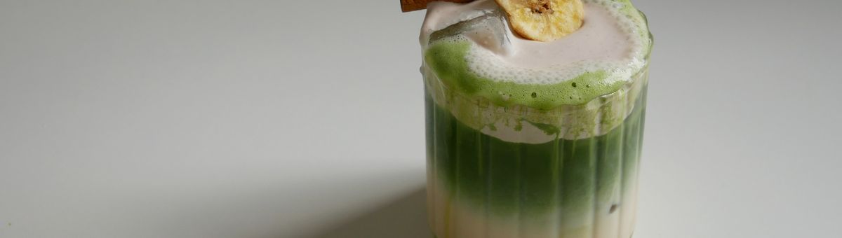 Ein Glas auf einem weißen Tisch, gefüllt mit einem Getränk. Unten ist eine milchige Flüssigkeit, darüber eine Schicht aus leuchtend grünem Matcha-Tee. Das Getränk ist mit Eiswürfeln versehen und hat eine Schaumkrone, die bis über den Rand läuft. Auf dem Schaum liegen eine Zimtstange und eine getrocknete Bananenscheibe.