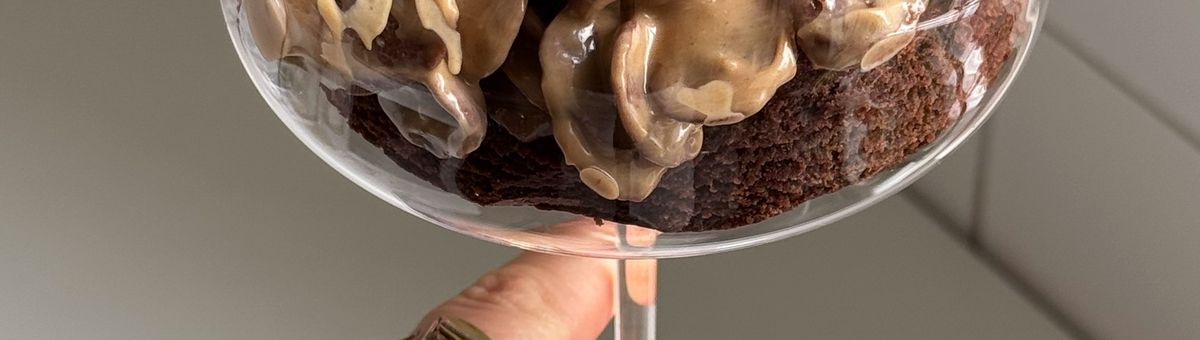 Ein Close-up-Foto zeigt einen dunklen Schokoladen-Brownie-Kuchen, der in einem eleganten Martiniglas serviert wird. Der Kuchenboden ist mit einer großzügigen Schicht einer cremigen, hellbraunen Masse bedeckt, in die knusprige, kakaofarbene Cerealien gemischt sind. Das Glas wird von unten von einer Hand mit einem goldenen Ring gehalten, die einen schwarzen Ärmel trägt.
