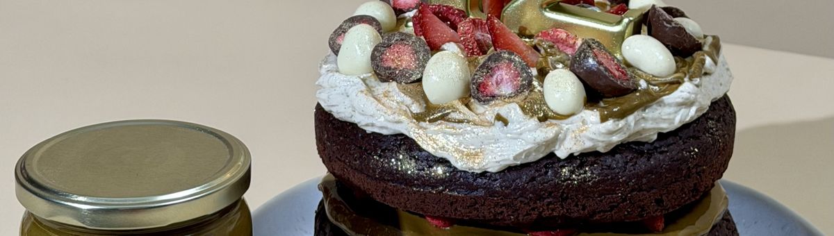 Veganer Schokoladen-Geburtstagskuchen mit goldenen Zahlenkerzen „12“, gefüllt mit Pistazienmus und frischen Erdbeeren, dekoriert mit Sahne, halbierten Schoko-Erdbeeren und weißen Schoko-Eiern auf einem hellen Teller; daneben ein Glas KoRo Pistazienmus – festlich dekorierte Geburtstagstorte zum 12. Geburtstag.
