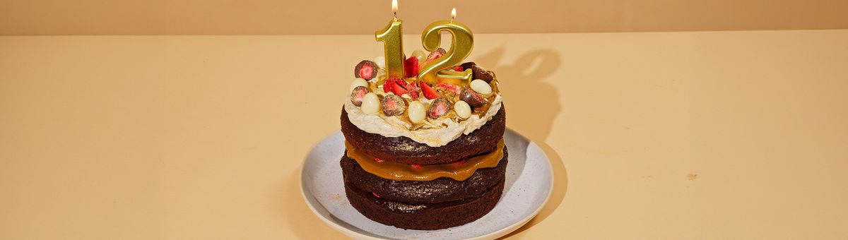 Veganer Schokoladen-Geburtstagskuchen mit goldenen Zahlenkerzen „12“, gefüllt mit Pistazienmus und frischen Erdbeeren, dekoriert mit Sahne, halbierten Schoko-Erdbeeren und weißen Schoko-Eiern auf einem hellen Teller; daneben ein Glas KoRo Pistazienmus – festlich dekorierte Geburtstagstorte zum 12. Geburtstag.