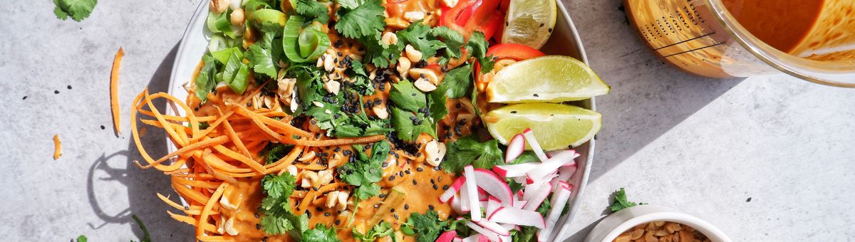 Thai Nudelsalat mit cremiger Erdnusssauce, frischem Gemüse, Koriander, Limettenspalten und gerösteten Erdnüssen in einer Schüssel