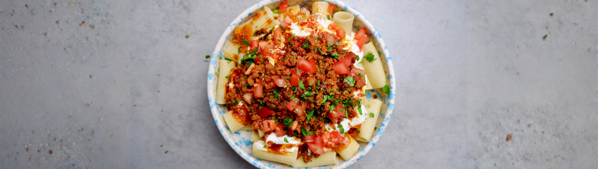 Ein Teller mit türkischer Pasta (Manti-Style): Große Rigatoni-Nudeln getoppt mit cremigem Knoblauchjoghurt, würzig angebratenem veganen Hackfleisch, frischen Tomatenwürfeln und gehackter Petersilie in einer blau-weiß gesprenkelten Schüssel.