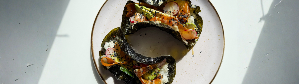 Zwei knusprige Sushi Tacos aus Nori-Blättern, gefüllt mit Sushi-Reis, Avocado, Radieschen, veganer Karotten-Lachs und schwarzer Sesam, angerichtet auf einem hellen Teller im Sonnenlicht.
