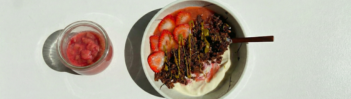 Veganes Erdbeer-Rhabarber-Kompott im Glas neben einer Bowl mit pflanzlichem Joghurt, frischen Erdbeeren, Schoko-Granola und Nussmus als Topping.