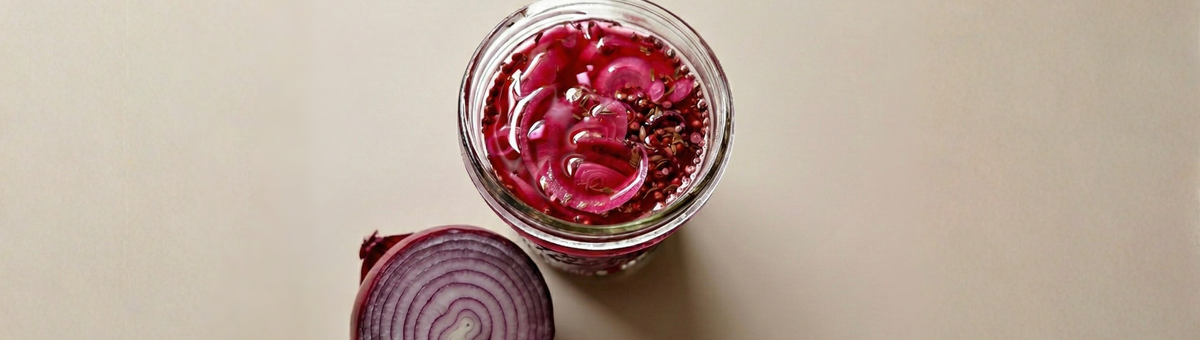 Glas mit pink eingelegten roten Zwiebelringen in Essig Sud mit Senfsamen und Koriandersamen, daneben eine halbierte rote Zwiebel auf hellem Hintergrund – selbstgemachte Pickled Onions.