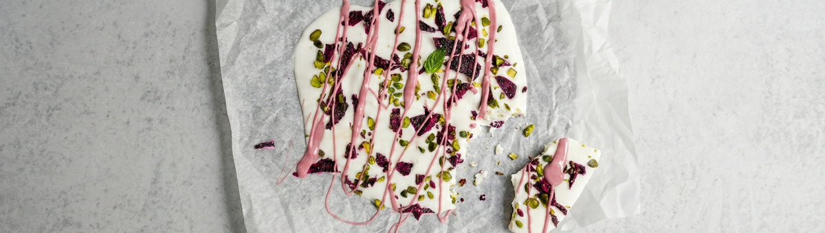 Gefrorener Joghurt auf einem mit Backpapier belegten Blech, getoppt mit getrockneten Drachenfruchtscheiben, Pistazien, frischer Minze und geschmolzener Ruby-Schokolade.