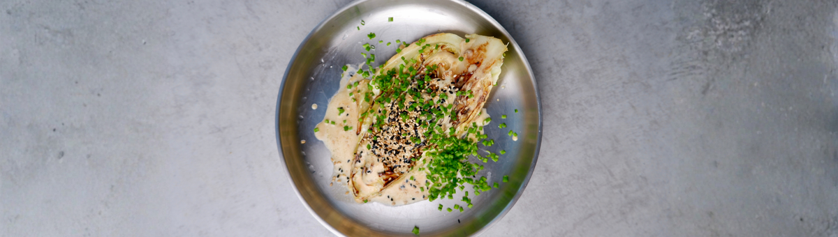 Gebratener Spitzkohl mit cremiger Miso-Rahm-Soße, garniert mit schwarzem und hellem Sesam sowie frischem Schnittlauch auf einem Metallteller angerichtet.