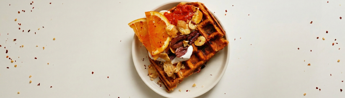 Herzhafte Orange-Chili-Waffel mit Orangenscheiben, veganem Joghurt, Oliven, Chili-Flocken und gerösteten Favabohnen auf einem weißen Teller.