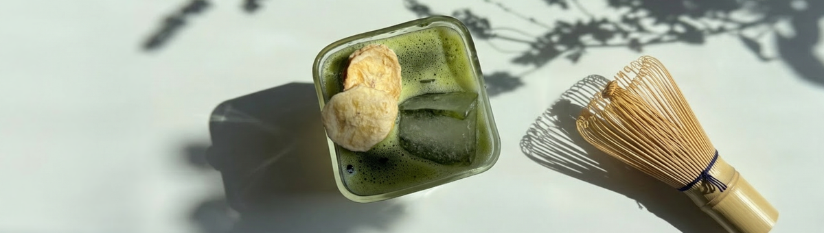 Ein eisgekühlter Matcha mit Kokoswasser und Bananenpulver, garniert mit getrockneten Bananenscheiben in einem quadratischen Glas, daneben ein traditioneller Matcha-Besen aus Bambus im Sonnenlicht.