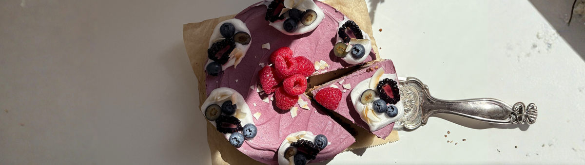 Draufsicht einer runden, lilafarbenen Cashew-Eistorte auf Backpapier. Ein schmuckvoller Tortenheber aus Silber hebt ein angeschnittenes Stück an. Die Torte ist mit Tupfen aus weißer Aquafaba-Sahne, frischen Heidelbeeren, Brombeeren, Himbeeren und Kokoschips garniert. Im Zentrum liegt ein Häufchen roter Himbeeren auf der cremig-gefrorenen Oberfläche.