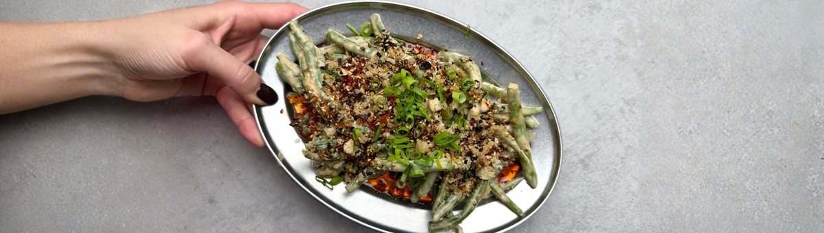 Grüne Bohnen mit cremiger Tahini-Sauce, Chili-Öl, Panko-Brösel, schwarzem Sesam und Frühlingszwiebeln auf einer Servierplatte.