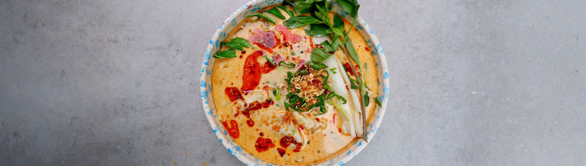 Schale mit cremiger Kokos-Curry-Dumpling-Suppe, gefüllt mit Gyoza und Pak Choi in aromatischer Brühe aus Kokosmilch, roter Currypaste, Ingwer und Knoblauch, garniert mit Frühlingszwiebeln, Röstzwiebeln, Chiliöl und frischen Kräutern auf hellem Hintergrund.