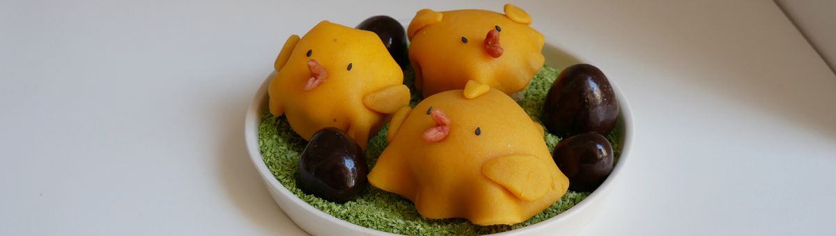 Drei kleine, runde Osterküken-Kuchen aus gelbem Marzipan mit rosa Schnäbeln und schwarzen Augen aus Sesam. Die Küken sitzen auf grün gefärbten Kokosraspeln in einer weißen Schale, dekoriert mit dunklen Schokoladeneiern auf weißem Hintergrund.