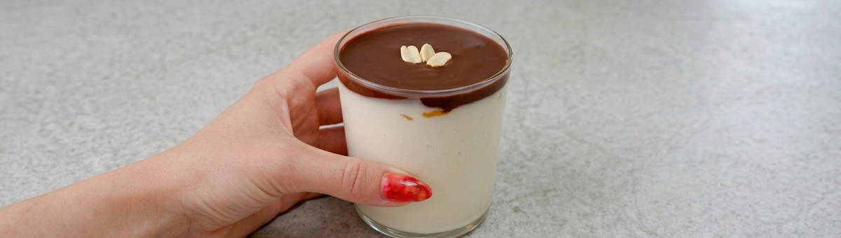 Ein cremiges Protein-Erdnuss-Dessert in einem Glas, serviert auf einer minimalistischen, grauen Arbeitsplatte. Das Dessert besteht aus einer hellen Vanille-Protein-Schicht, getoppt mit einer glatten, dunklen Schokoladenglasur und garniert mit drei Erdnüssen in der Mitte. Eine Hand mit rot lackierten Fingernägeln hält das Glas seitlich fest.