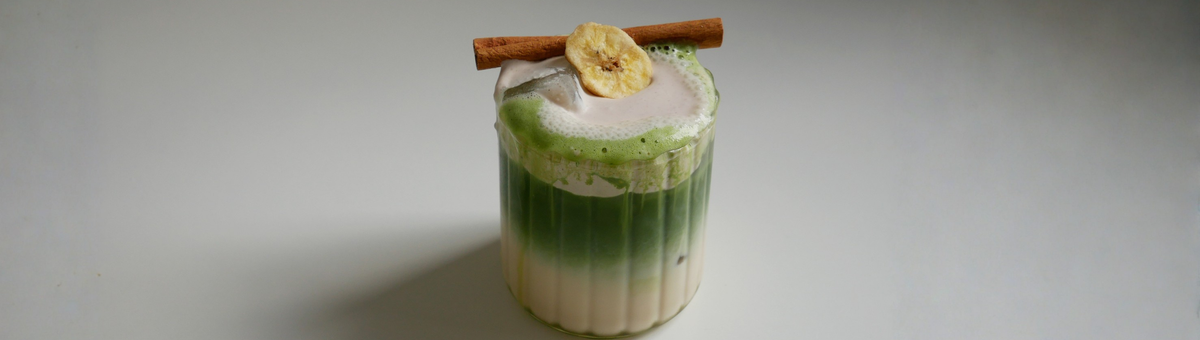 Ein Glas auf einem weißen Tisch, gefüllt mit einem Getränk. Unten ist eine milchige Flüssigkeit, darüber eine Schicht aus leuchtend grünem Matcha-Tee. Das Getränk ist mit Eiswürfeln versehen und hat eine Schaumkrone, die bis über den Rand läuft. Auf dem Schaum liegen eine Zimtstange und eine getrocknete Bananenscheibe.