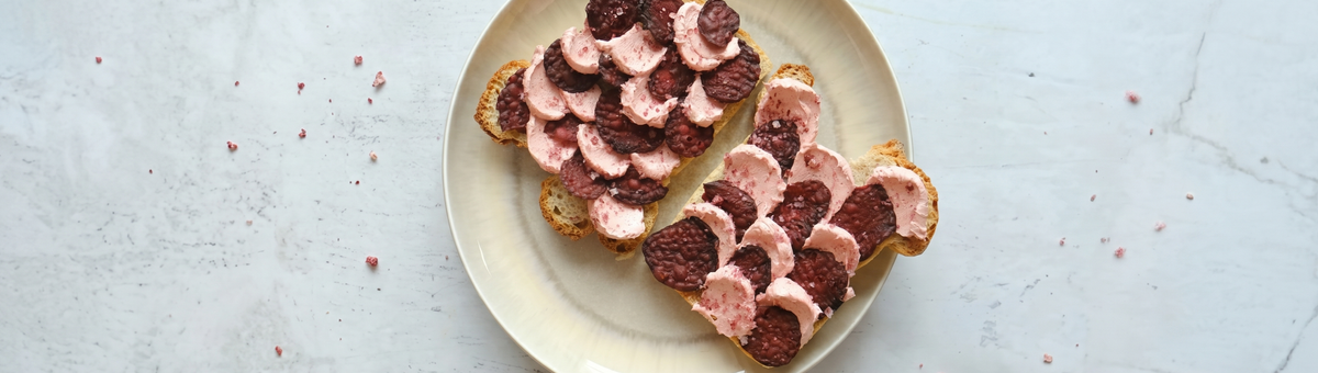 Zwei Scheiben geröstetes Brot auf einem hellen Teller, belegt mit rosa aufgeschlagener veganer Butter und dunklen Rote-Bete-Chips in einem dekorativen Schuppenmuster auf einer Marmoroberfläche.