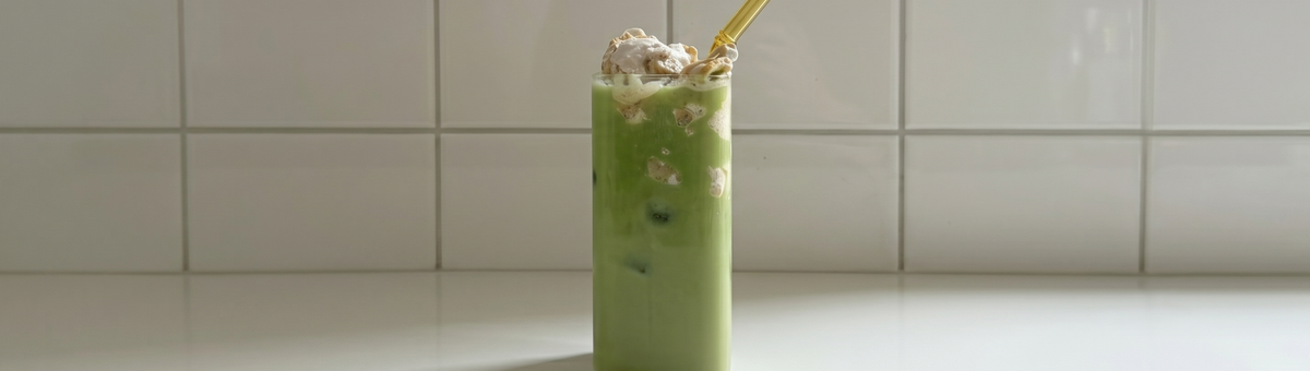 Ein hohes Glas mit grünem Matcha-Bananen-Pudding Drink und gelbem Strohhalm vor einer weißen Fliesenwand.