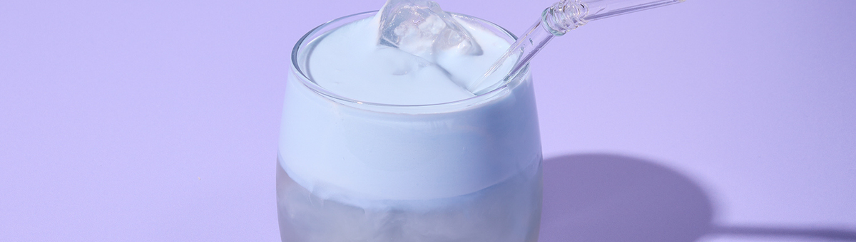 Cremiger Blue Spirulina Coconut Cloud Drink mit Eiswürfeln und Glasstrohhalm vor lila Hintergrund – modernes Trendgetränk.