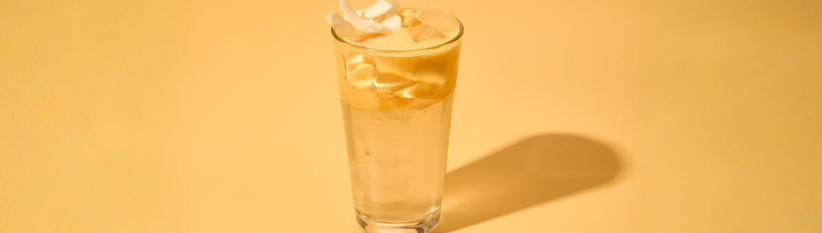 Erfrischender Mango Coconut Cloud Water Drink mit Eiswürfeln und Kokoschips in einem Glas vor gelbem Hintergrund – tropisches Sommergetränk.