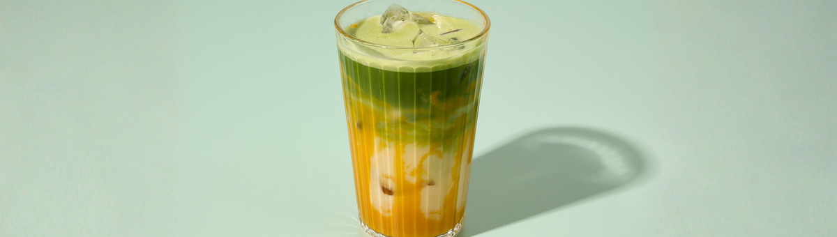 Grüner Matcha-Mango Latte mit Milch und Eiswürfeln im Glas vor mintfarbenem Hintergrund – iced Matcha-Getränk in hochwertiger Produktfotografie.