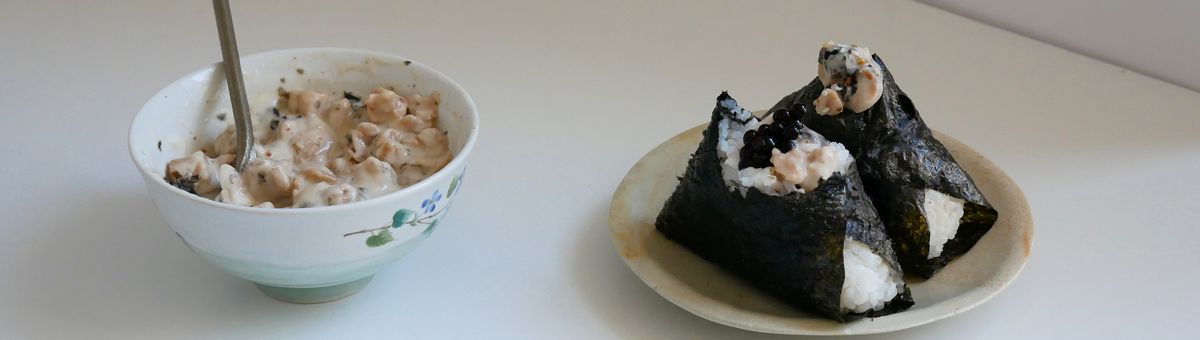Zwei vegane Tuna-Mayo-Onigiri umwickelt mit Nori-Alge auf einem Teller neben einer Schüssel mit veganer Thunfisch-Füllung.