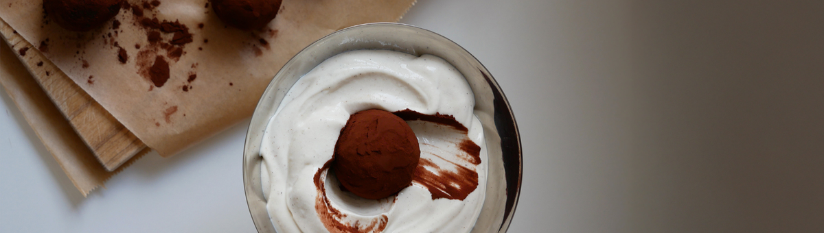 Tiramisu Energy Balls: Ein mit Kakaopulver bestäubtes Energiebällchen liegt eingebettet in cremiger weißer Creme in einer silbernen Schale, weitere Bällchen im Hintergrund auf Backpapier.