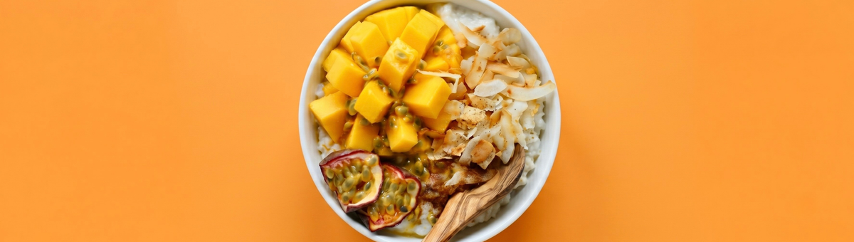 Cremige Frühstücks-Bowl mit Milchreis, frischen Mangowürfeln, Passionsfrucht und gerösteten Kokoschips mit einem Holzlöffel auf hellorangem Hintergrund.