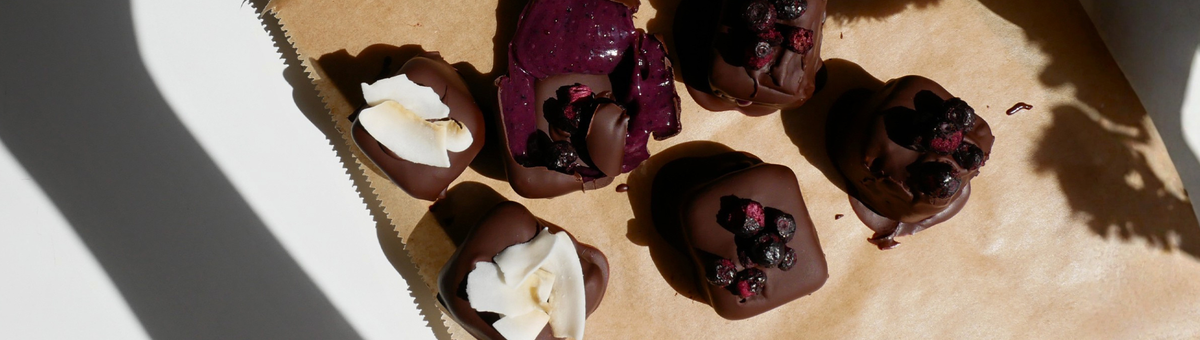 Draufsicht auf fünf Mango-Matcha-Ube-Eis-Bites, überzogen mit Schokolade und garniert mit Kokosraspeln und Johannisbeeren, arrangiert auf Backpapier im Sonnenlicht.