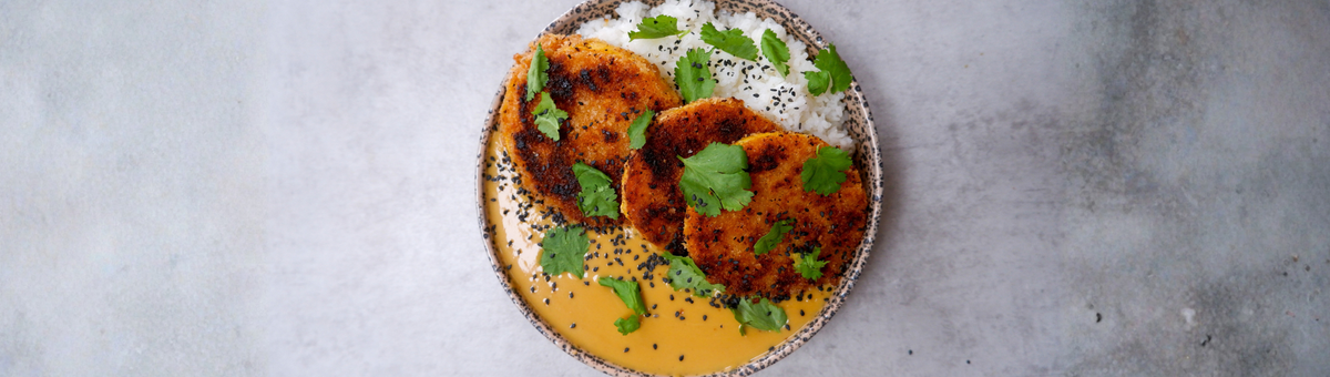 Zentrierte Draufsicht auf eine gesprenkelte Schüssel mit knusprig panierten Butternut-Kürbis-Scheiben, serviert mit weißem Reis und einer cremigen, gelborangen Currysauce. Das Gericht ist mit frischen Korianderblättern und schwarzem Sesam garniert und steht auf einem schlichten, hellgrauen, leicht strukturierten Untergrund im Querformat.
