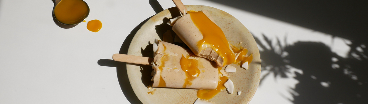 Zwei vegane Mango-Vanille Popsicles auf einem rustikalen Teller in hellem Sonnenlicht, großzügig übergossen mit fruchtigem Mangopüree und garniert mit gerösteten Kokoschips.