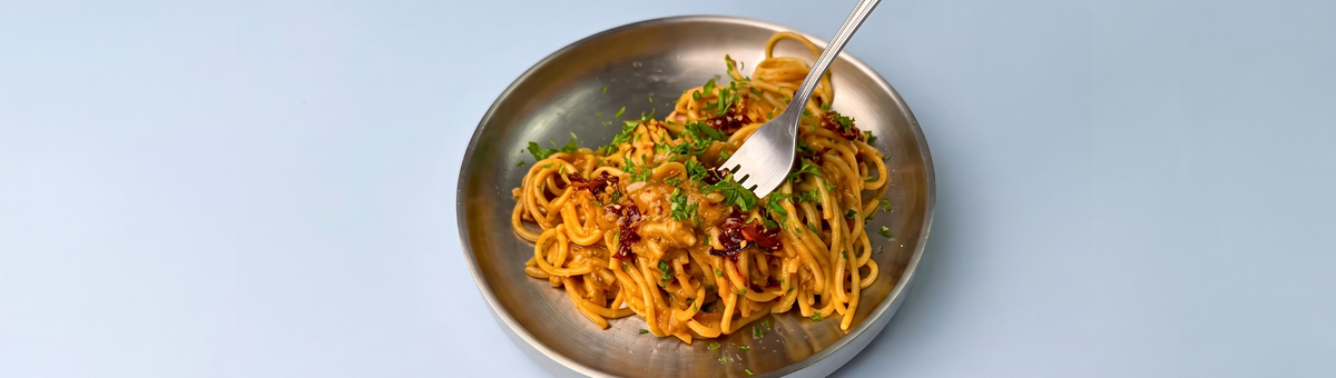 Ein Metallteller mit veganer Pasta mit karamellisierten Zwiebeln, garniert mit frischen Kräutern und getrockneten Tomatenstücken, in die eine Kuchengabel hineinsticht, fotografiert vor einem hellblauen Hintergrund.Ein Metallteller mit veganer Pasta mit karamellisierten Zwiebeln, garniert mit frischen Kräutern und getrockneten Tomatenstücken, in die eine Kuchengabel hineinsticht, fotografiert vor einem hellblauen Hintergrund.