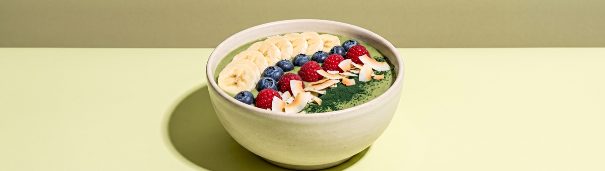 Eine hellgrüne Spriulina-Bowl in einer beigen Keramikschüssel, ordentlich garniert mit Bananenscheiben, Blaubeeren, Himbeeren und Kokoschips vor einem minimalistischen, grünen Hintergrund.