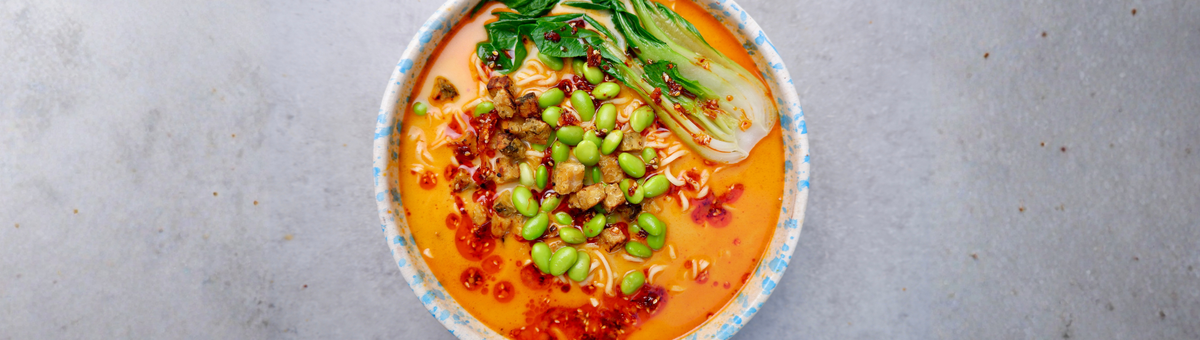 charfe Kokos-Ramen mit gebratenem Tempeh, grünem Gemüse und Chili-Öl in einer gesprenkelten Keramikschüssel.