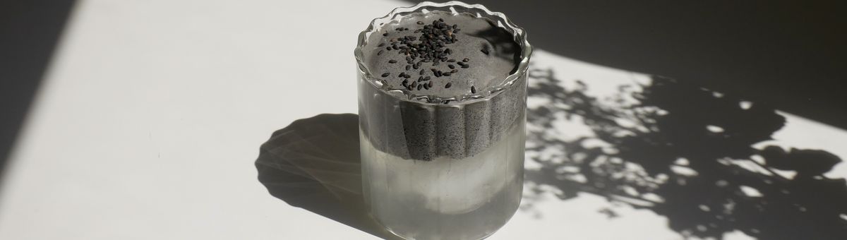 Schwarzer Sesam Cloud Latte in einem geriffelten Glas. Trendiges, geschichtetes Kaffee-Alternative-Getränk mit samtiger schwarzer Sesam-Creme und gerösteten Sesamsamen-Topping. Minimalistischer Stil mit natürlichem Sonnenlicht und Schattenspiel.