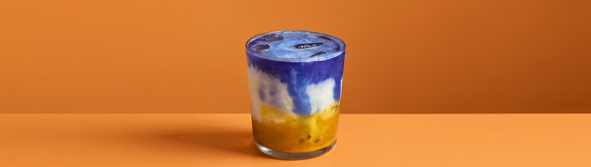Ein geschichteter Mango-Passionsfrucht-Blue-Spirulina-Latte in einem klaren Glas auf einem warmen, orangefarbenen Hintergrund.